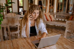 Woman sitting at wooden table, using laptop and talking on phone in a cozy indoor setting.