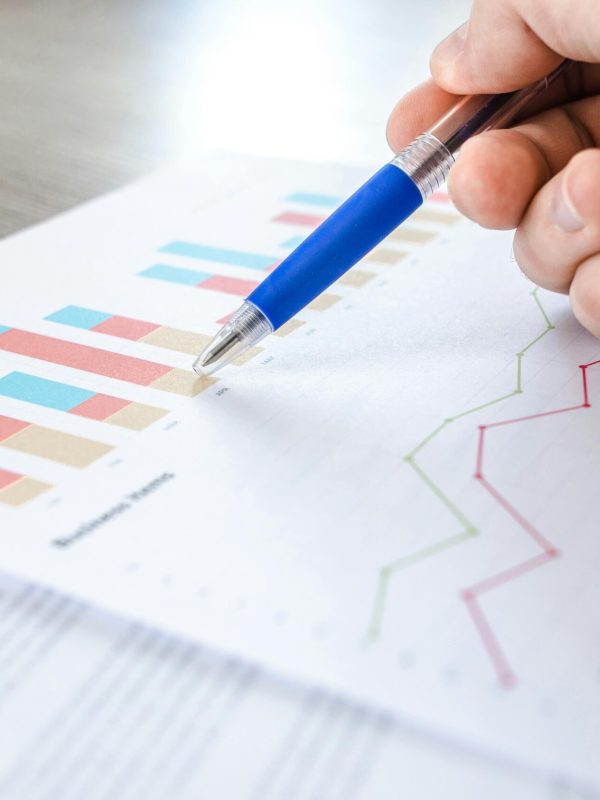 A close-up of a hand with a pen analyzing data on colorful bar and line charts on paper.